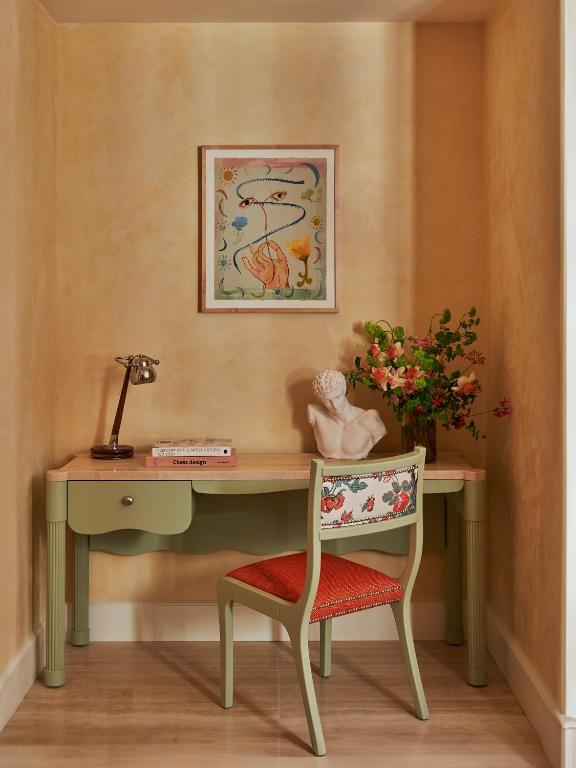 13 UNIQUE Boutique Hotels in Rome 43 A small, stylish desk nook reminiscent of boutique hotels in Rome, with a green desk and floral-accented chair. The desk holds books, a vintage phone, a white bust, and fresh flowers beneath whimsical framed artwork.
