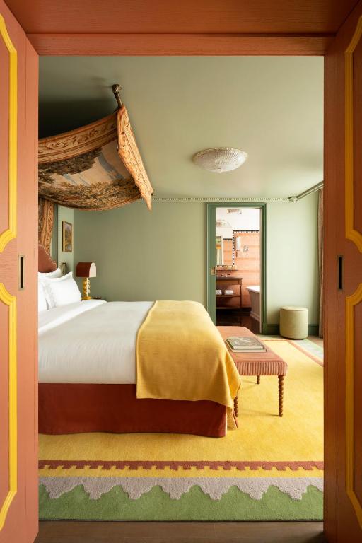 13 Unique Hotels in Paris for a Stylish Stay 37 A cozy hotel room with a canopy bed, yellow blanket, and patterned rug. Soft green walls, bedside lamp, and a bench at the foot of the bed create the charm found in unique hotels in Paris. A doorway leads to a peach-colored bathroom.