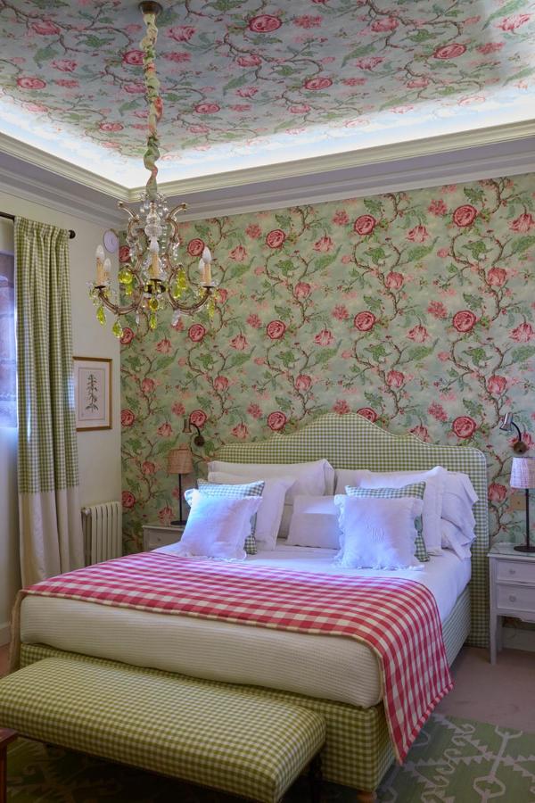 A cozy bedroom in the style of charming hotels in Cantabria, with floral wallpaper and matching ceiling, a bed with white pillows, green checkered headboard and bench, pink checkered throw, bedside tables, a chandelier, and patterned curtains.