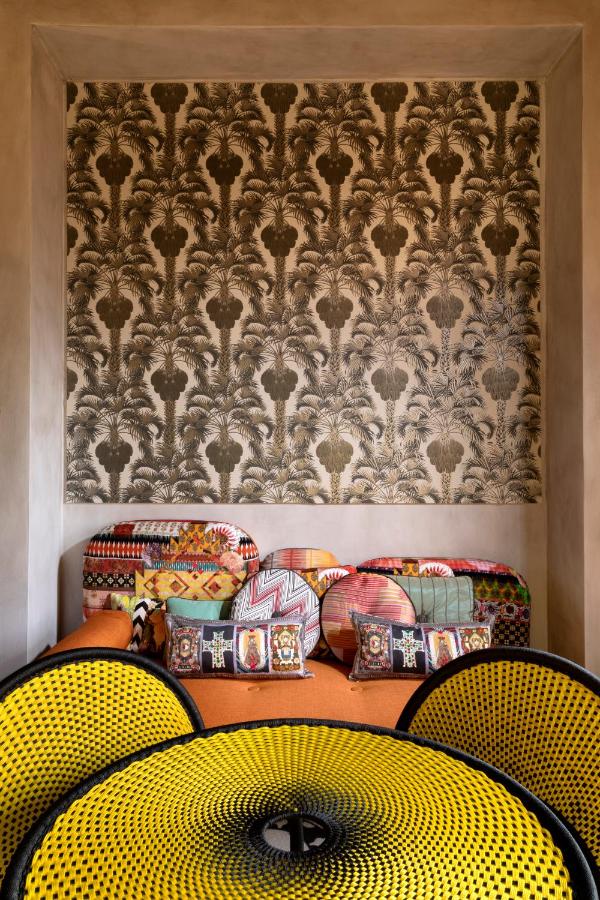 13 UNIQUE Boutique Hotels in Rome 38 A cozy seating area with colorful patterned cushions, a warm orange sofa, and bold yellow-and-black woven chairs evokes the inviting charm of boutique hotels in Rome, set against intricate palm tree-patterned wallpaper.
