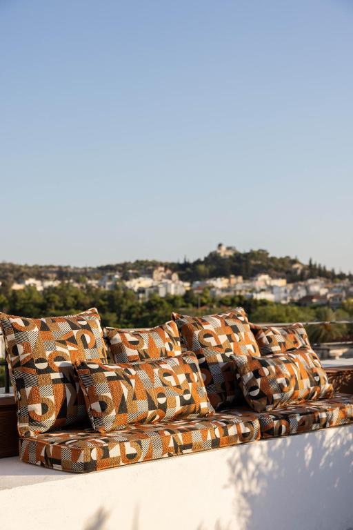 A bench with orange, brown, and white geometric-patterned cushions sits on a terrace—much like those in boutique hotels in Athens—overlooking a scenic town with hills and greenery under a clear blue sky.
