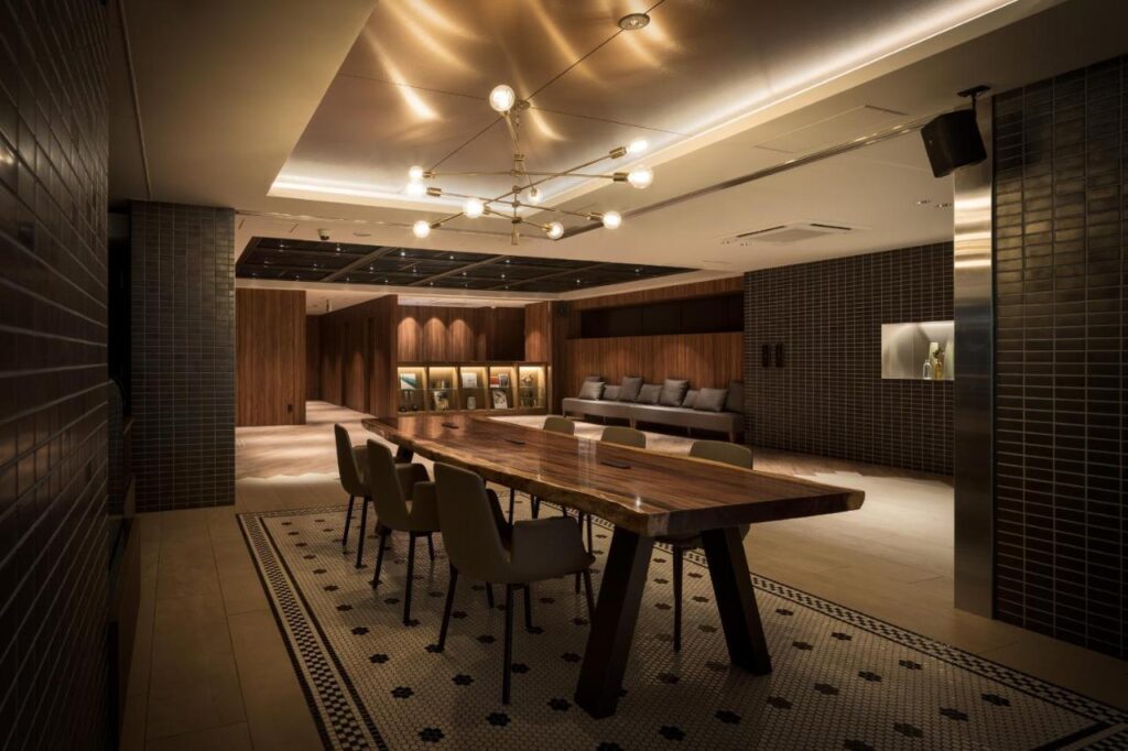 7 Best Hotels in Hiroshima, Japan 15 A modern, dimly-lit conference room in one of the best hotels in Hiroshima, Japan, featuring a long wooden table, several chairs, a sofa in the back, dark tiled walls, a geometric chandelier above, and bottles displayed on a recessed shelf.