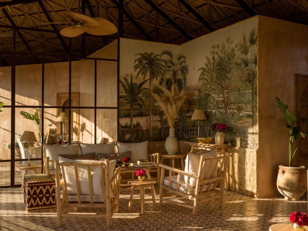 10 Unique Riads in Marrakech with Pool 20 A cozy room with bamboo furniture, neutral cushions, patterned tile floor, and a tropical mural captures the serene charm of riads in Marrakech with pool. Sunlight streams in, highlighting potted plants and decorative vases with red and white flowers.
