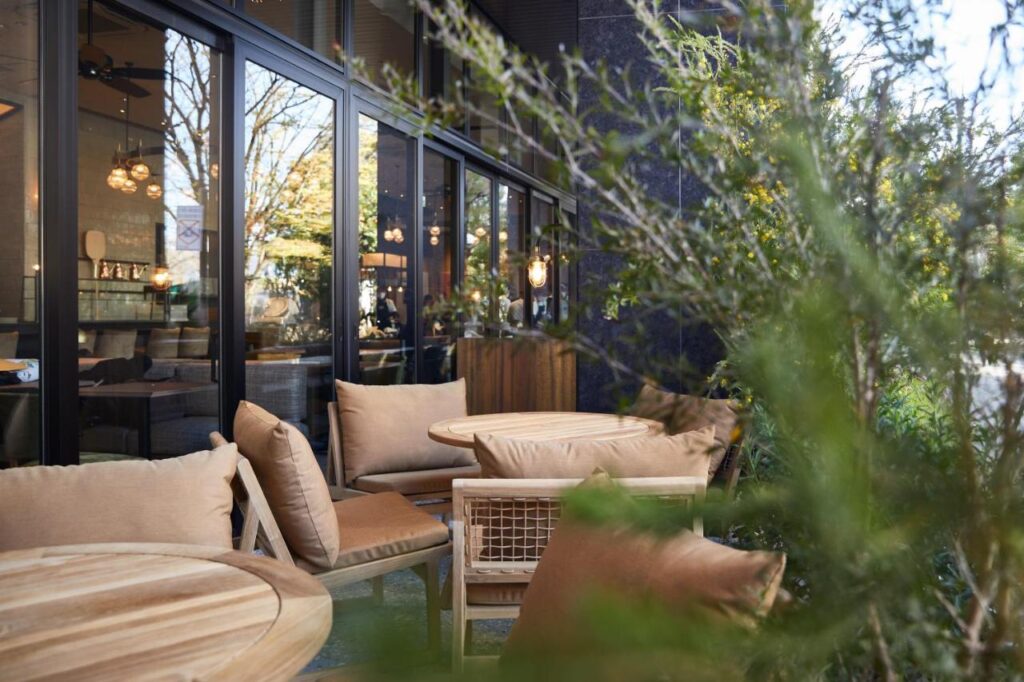 7 Best Hotels in Hiroshima, Japan 12 Outdoor seating area of a café or restaurant with round wooden tables and cushioned chairs—much like those at the best hotels in Hiroshima, Japan. Large windows reveal a cozy interior with warm lighting, while green plants partially obscure the foreground.