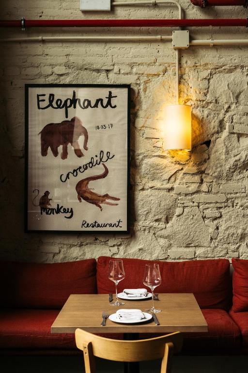 13 Unique Hotels in Barcelona, Spain 50 A cozy dining area in one of the unique hotels in Barcelona, featuring a wooden table set for two, red cushioned seating, exposed brick walls, a wall lamp, and a framed poster with elephant, crocodile, and monkey drawings.