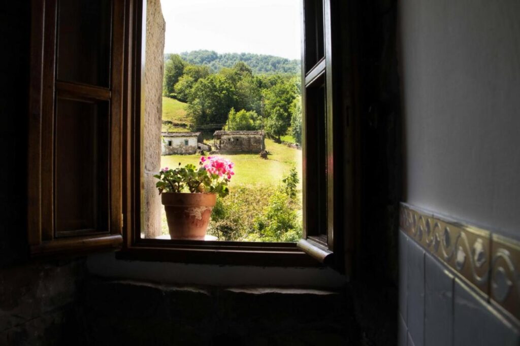 A potted pink flower sits on a windowsill, with the window open to reveal a green, sunlit countryside reminiscent of views from hotels in Cantabria—trees, grassy fields, and a distant farmhouse beyond the tiled wall and window frame.