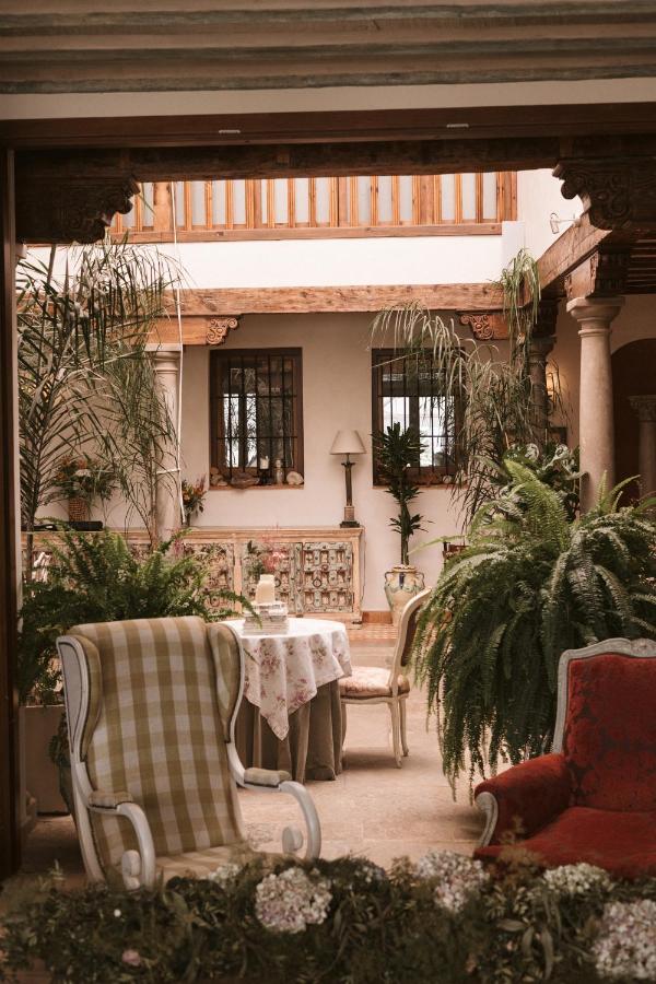 A cozy indoor courtyard reminiscent of boutique hotels in Granada, featuring vintage chairs, a floral tablecloth, lush green plants, and wooden accents—an inviting space filled with natural light.