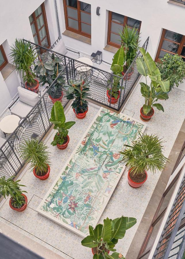 A courtyard viewed from above features a rectangular mosaic table surrounded by potted tropical plants—an inviting scene reminiscent of boutique hotels in Granada. The space is bordered by black railings and white walls with large windows.
