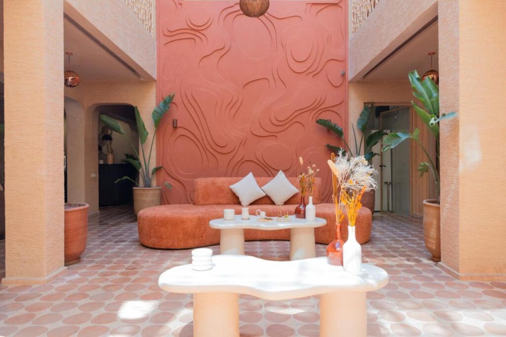 10 Unique Riads in Marrakech with Pool 23 A modern, sunlit lounge with peach and terracotta tones draws design inspiration from riads in Marrakech with pool, featuring a curved sofa, two white round tables with vases, patterned tile floor, abstract wall art, and lush potted plants.