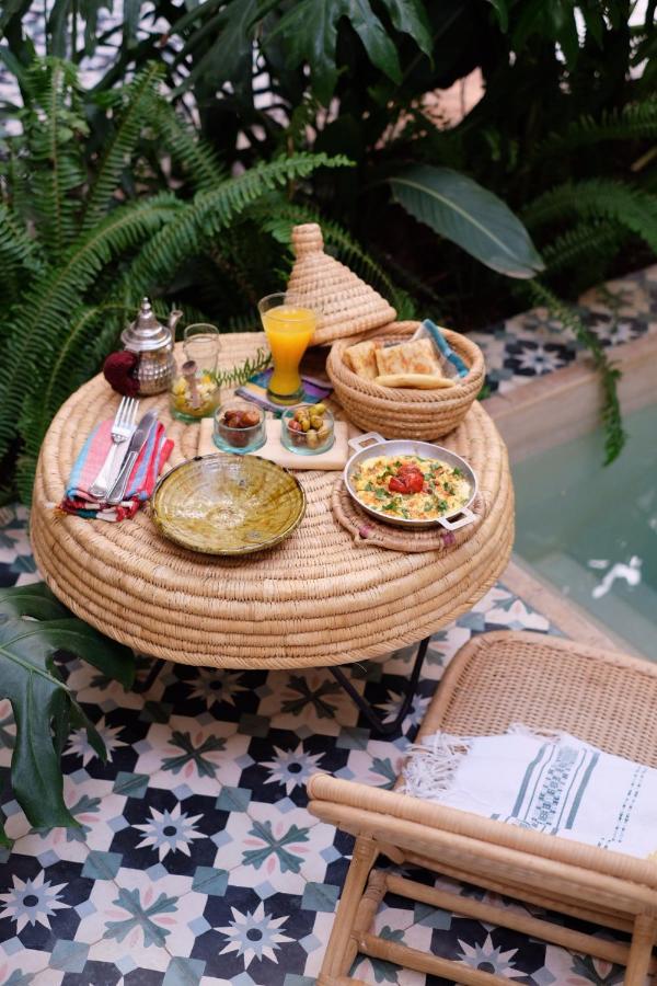 10 Unique Riads in Marrakech with Pool 34 A round wicker table set with Moroccan dishes, orange juice, bread, and tea sits beside a pool, surrounded by lush greenery and patterned tiles—capturing the serene charm of riads in Marrakech with pool.