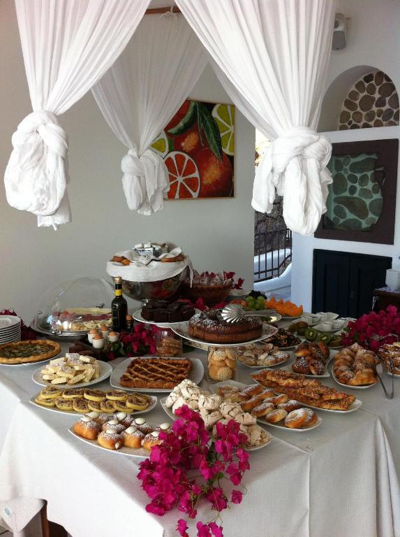 7 Best Hotels in the Aeolian Islands 24 A buffet table covered with a white cloth holds a variety of pastries, cakes, and breads, adorned with pink flowers—evoking the elegant breakfast spreads found in hotels in the Aeolian Islands. White drapes hang above, with colorful fruit artwork on the wall behind.