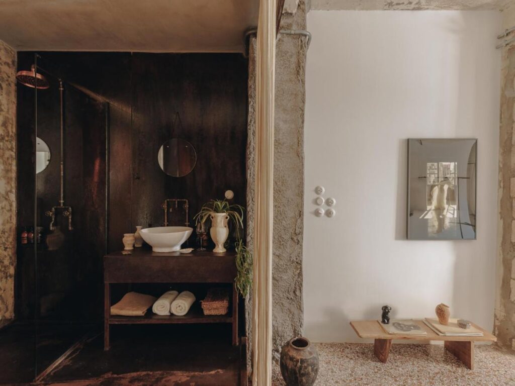 A rustic bathroom reminiscent of the best boutique hotels in Athens features a dark, industrial-style sink with a round mirror and white basin, while an adjoining space showcases a wooden bench, vases, wall art, and a large mirror on a white wall.