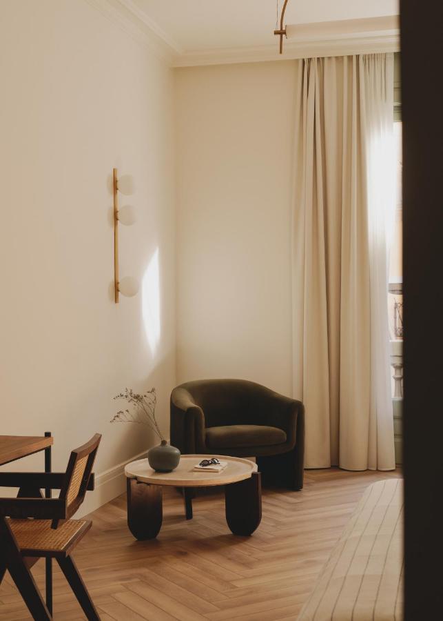 13 Unique Hotels in Barcelona, Spain 16 A cozy minimalist living room with a dark green armchair, round coffee table with a vase, wooden chairs, light beige walls, and tall cream curtains letting in natural sunlight—like the inviting spaces found in unique hotels in Barcelona.