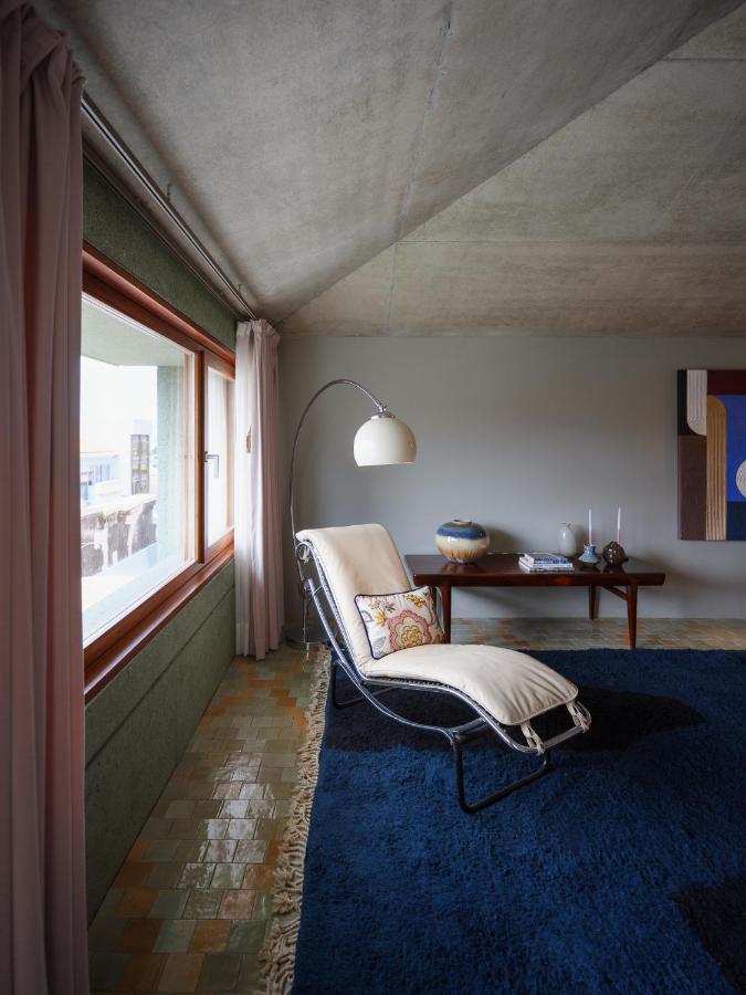 Top 12 Boutique Hotels in Porto, Portugal 18 A cozy lounge chair with a pillow and blanket sits by a large window with pink curtains, evoking the charm of boutique hotels in Porto. A modern floor lamp arches overhead, while a blue rug and stylish side table complete the serene room.