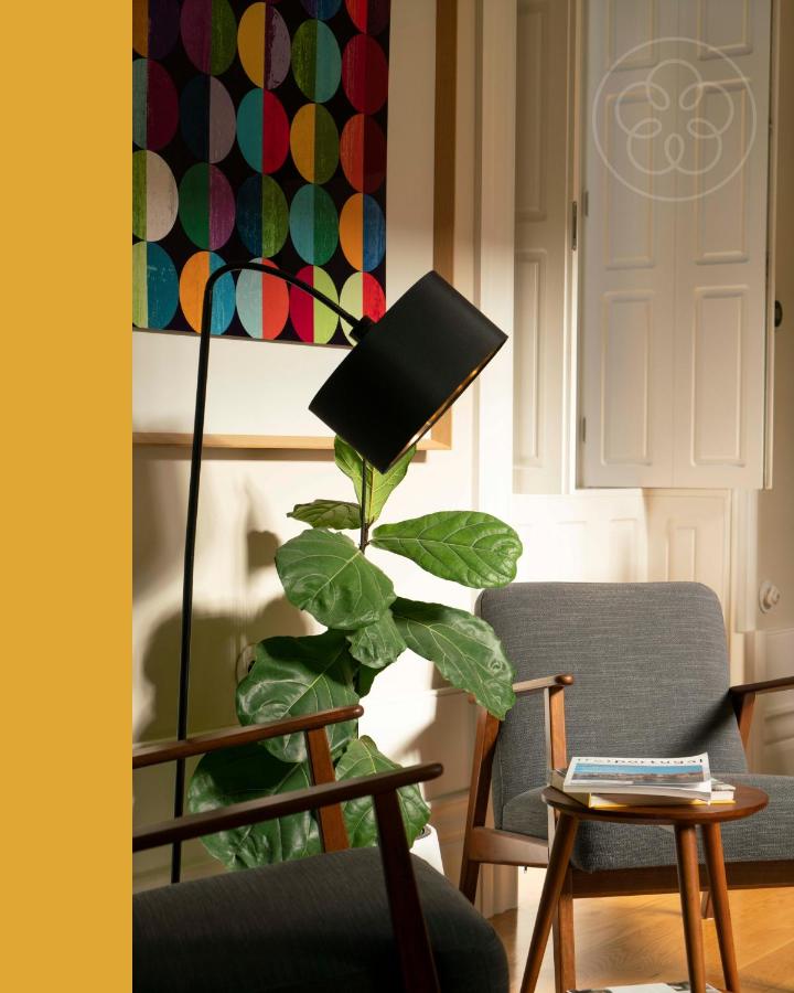 Top 12 Boutique Hotels in Porto, Portugal 14 A cozy living room corner inspired by boutique hotels in Porto, featuring a dark gray armchair, a small wooden side table with magazines, a leafy potted plant, a black floor lamp, and colorful geometric artwork on the wall.