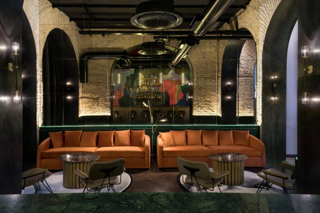 13 UNIQUE Boutique Hotels in Rome 9 Modern lounge with exposed brick arches and industrial ceiling features. Two orange sofas face three round tables with chairs. Reminiscent of boutique hotels in Rome, Italy, a colorful mural and bar glow in the softly lit background.