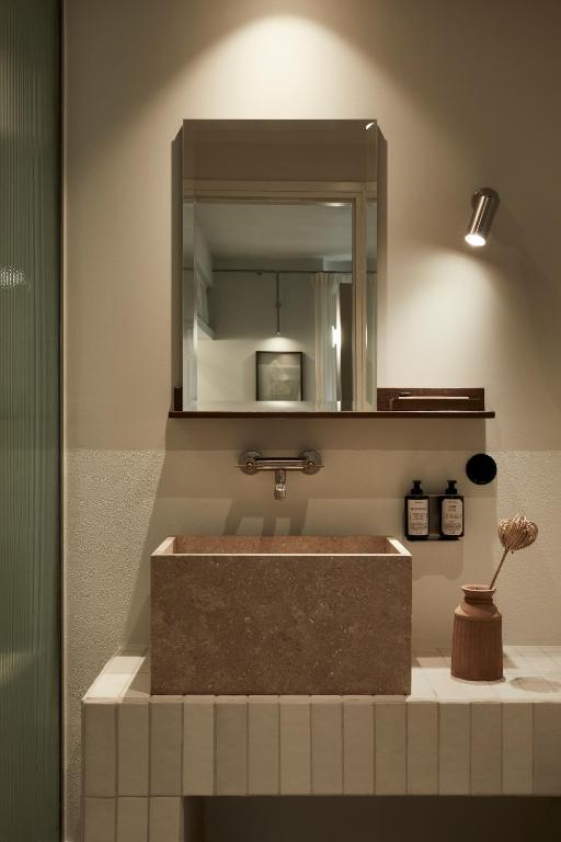 A modern bathroom inspired by boutique hotels in Athens features a rectangular stone sink, wall-mounted faucet, and mirror. Two soap dispensers and wooden decor sit on the tiled countertop, illuminated by a wall spotlight above.