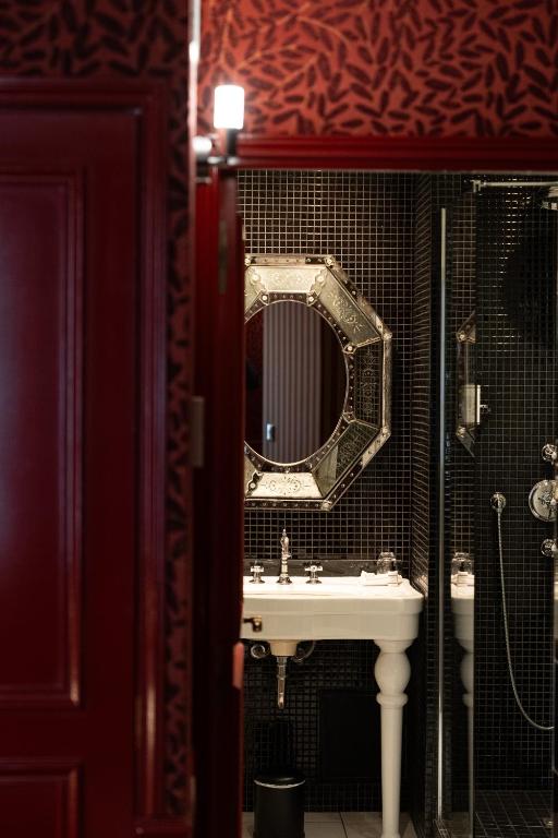 13 Unique Hotels in Paris for a Stylish Stay 51 A view through a doorway into a bathroom with black tiled walls, a round decorative mirror, a white pedestal sink, and a glass shower; the door and wall are dark red—evoking the stylish charm of unique hotels in Paris.