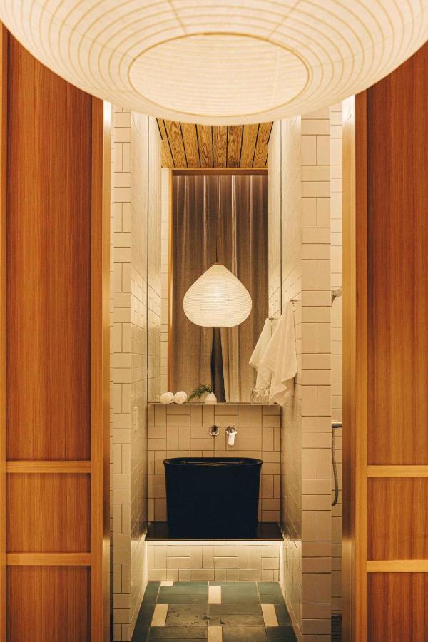 15 Unique Hotels in Tokyo: Coolest hotels 12 A modern bathroom in the style of unique hotels in Tokyo, featuring white tiled walls, a black soaking tub, a hanging round paper lantern, wooden accents, and soft, warm lighting for a cozy atmosphere.