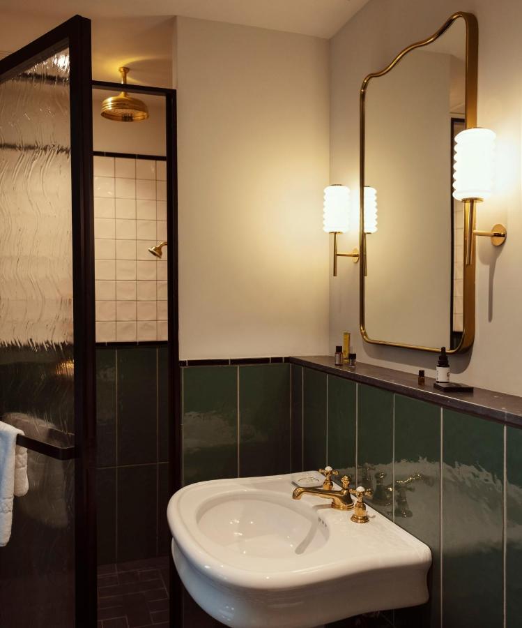 13 UNIQUE Boutique Hotels in Rome 27 A stylish bathroom inspired by boutique hotels in Rome, Italy, features green and white tiles, a white sink with gold fixtures, a gold-framed mirror, wall-mounted lights, and a shower with a glass door and gold rain showerhead.
