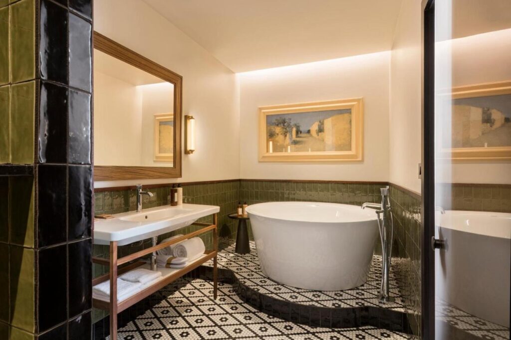 13 Unique Hotels in Paris for a Stylish Stay 22 A modern bathroom, reminiscent of unique hotels in Paris, features a freestanding white bathtub, double-sink vanity with towels beneath, green tiled walls, patterned black-and-white floor tiles, a large mirror, and framed artwork on the wall.