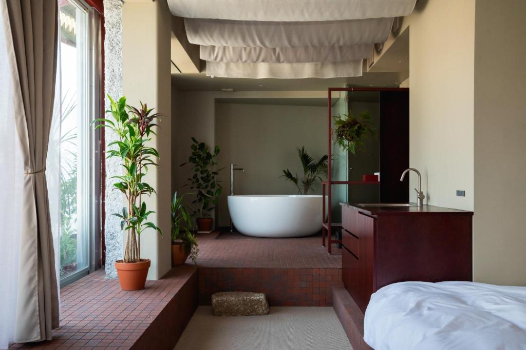 15 Unique Hotels in Tokyo: Coolest hotels 40 A modern bathroom inspired by unique hotels in Tokyo features a freestanding white tub, potted plants, a sink with a wooden cabinet, and large windows with curtains letting in natural light.
