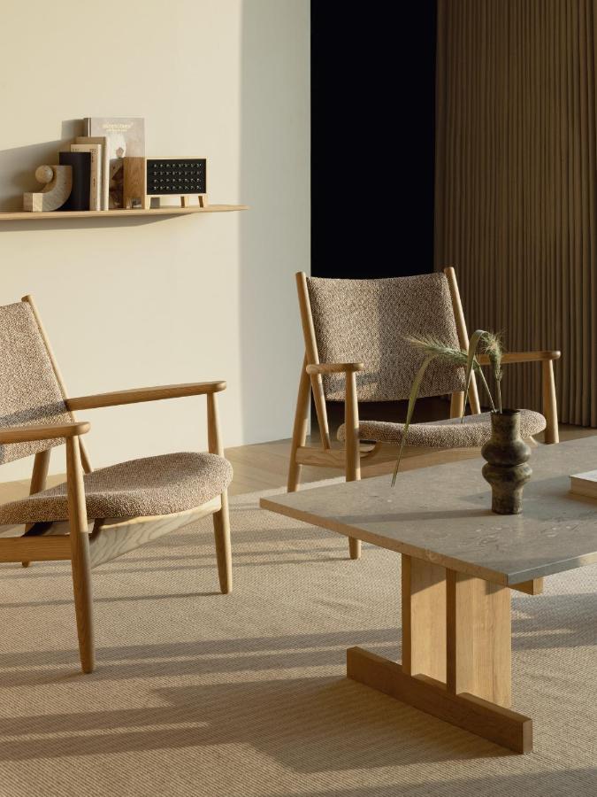 15 Unique Hotels in Tokyo: Coolest hotels 2 Two wooden armchairs with textured beige cushions sit near a light wood coffee table with a decorative vase, evoking the serene style found in unique hotels in Tokyo. Minimalist decor and books line a floating shelf, while natural light fills the space.
