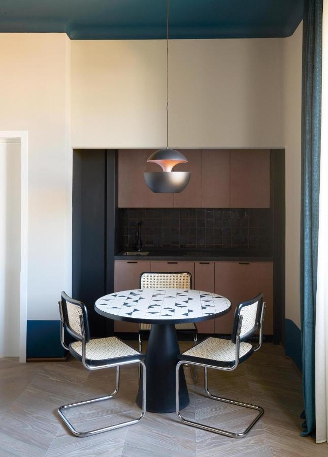 13 UNIQUE Boutique Hotels in Rome 16 A modern dining nook with a round table and three chairs sits in front of a small kitchen—reminiscent of those found in boutique hotels in Rome, Italy. A spherical pendant light hangs above, with sunlight streaming through curtained windows.