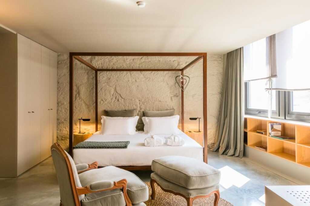 Top 12 Boutique Hotels in Porto, Portugal 22 A bright, modern bedroom reminiscent of boutique hotels in Porto, featuring a four-poster bed with white bedding, two armchairs with ottomans, a stone wall, large windows with curtains, and a wooden bookshelf. Towels are neatly placed on the bed.