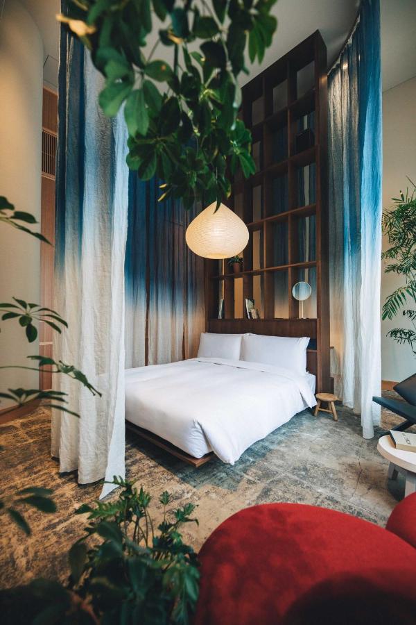 15 Unique Hotels in Tokyo: Coolest hotels 10 A modern bedroom with a low, white bed, tall wooden shelving, and blue-and-white gradient curtains—like those found in unique hotels in Tokyo. Green plants and a round paper lantern add a fresh, natural touch to the cozy, stylish space.