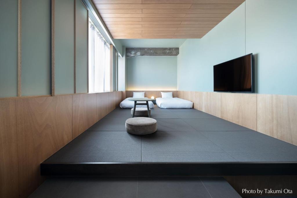 15 Unique Hotels in Tokyo: Coolest hotels 47 A minimalist Japanese-style room in one of the unique hotels in Tokyo, featuring two futon beds, a low table with a round cushion, wooden wall panels, large windows, and a wall-mounted TV. Natural light fills the clean, open space.