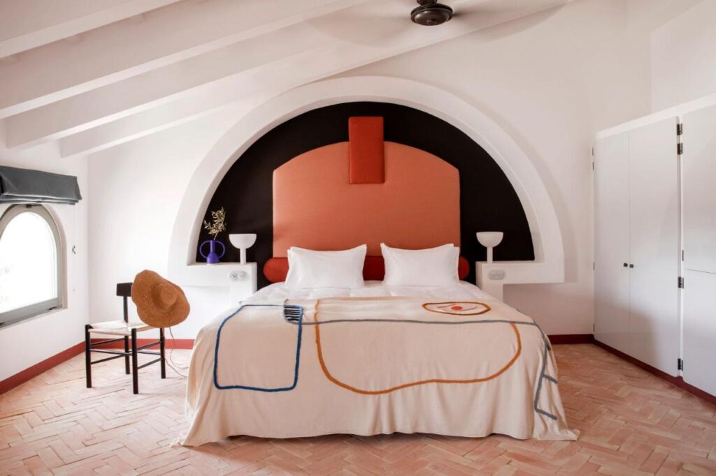 Unique Boutique Hotels in Menorca 24 A stylish bedroom inspired by boutique hotels in Menorca, featuring a double bed with a white and orange headboard, patterned blanket, two white pillows, sleek white lamps, a black chair with a tan hat, and built-in closets under a slanted ceiling.