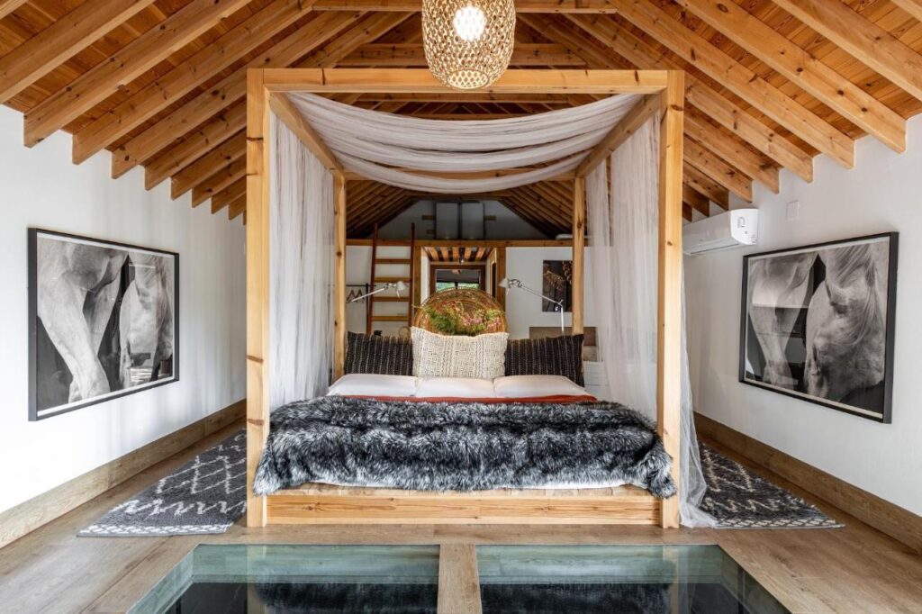 A cozy bedroom inspired by hotels in Cantabria, with a wooden canopy bed, draped white curtains, fur throw, and decorative pillows. Exposed wood beam ceiling, white walls, and black-and-white horse photos complete the serene retreat.