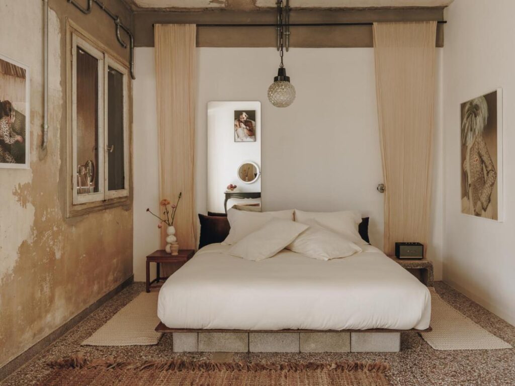 A cozy bedroom with a minimalist design, inspired by the best boutique hotels in Athens, features a double bed with white bedding, pillows, and a wooden side table adorned with vases and dried flowers. Photos decorate the walls, and a pendant light hangs above the bed.