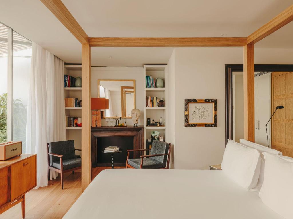 13 Unique Hotels in Barcelona, Spain 26 A bright, modern bedroom in one of the unique hotels in Barcelona features a four-poster bed, white linens, wooden furniture, bookshelves, and a large mirror above a dark fireplace. Soft natural light streams in from sheer-curtained windows.