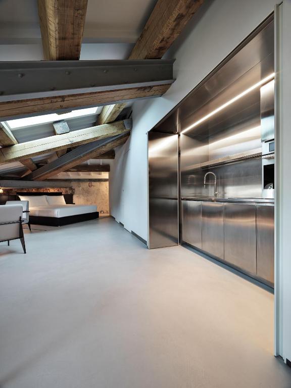 13 UNIQUE Boutique Hotels in Rome 32 Modern loft reminiscent of boutique hotels in Rome, featuring exposed wooden beams, stainless steel kitchen appliances, minimalist cabinets, light floors, and an open layout flowing to a cozy living area with a couch and bed beneath slanted ceiling windows.