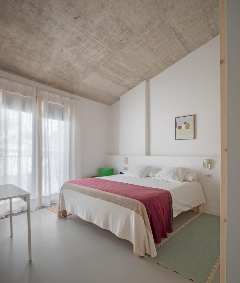 Unique Boutique Hotels in Menorca 35 Minimalist bedroom inspired by boutique hotels in Menorca, featuring a concrete ceiling, white walls, a large bed with white linens and a red blanket, sheer curtains over glass doors, a small desk, and simple wall art above the bed.