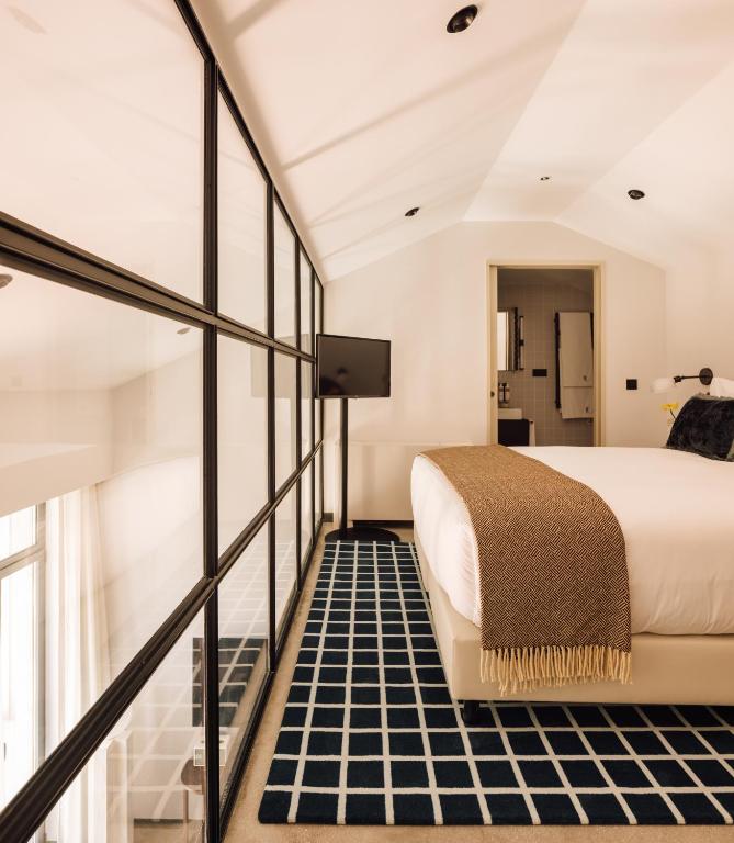 Top 12 Boutique Hotels in Porto, Portugal 35 Modern bedroom with a large glass wall, a neatly made bed with a brown throw, black and white checkered rug, TV on a stand, and an open doorway to the bathroom—reminiscent of stylish boutique hotels in Porto.