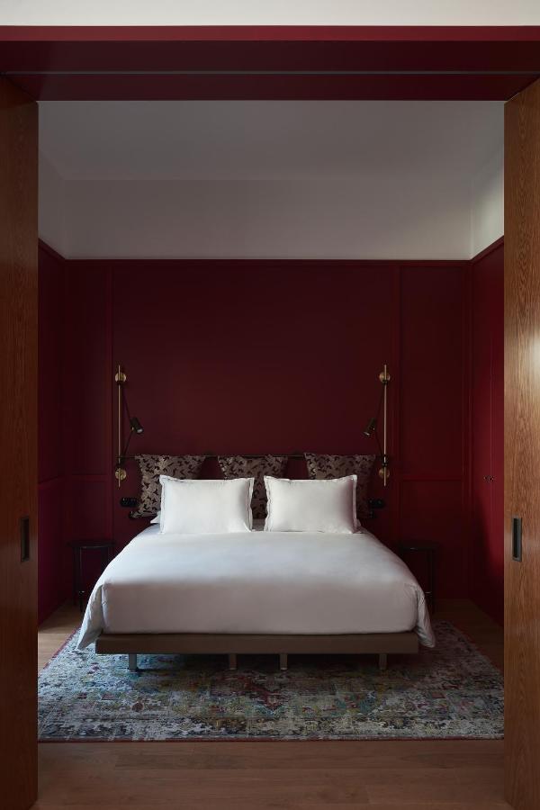 13 Unique Hotels in Paris for a Stylish Stay 17 A modern bedroom inspired by unique hotels in Paris, featuring deep red walls, a bed with white linens and patterned pillows, wall-mounted reading lamps, and a colorful rug on a light wooden floor framed by wooden sliding doors.