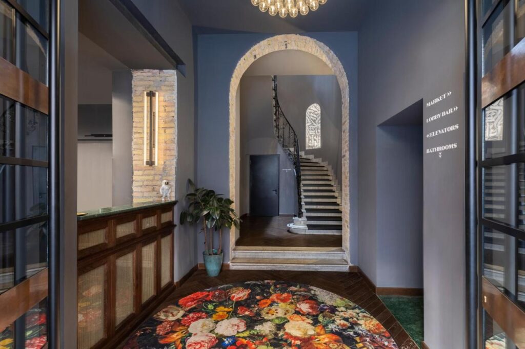 13 UNIQUE Boutique Hotels in Rome 10 A stylish entryway reminiscent of boutique hotels in Rome, Italy, features a floral rug, arched brick doorway, marble staircase with wrought iron railing, and modern lighting. A potted plant sits near the arch with directions displayed on the wall.