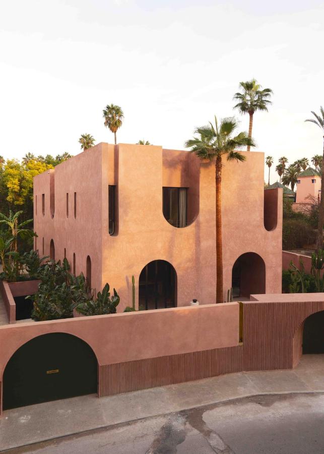 10 Unique Riads in Marrakech with Pool 9 A modern, pink-toned, two-story building with arched doorways and windows evokes the elegance of riads in Marrakech with pool, surrounded by lush greenery and tall palm trees beneath a bright sky.