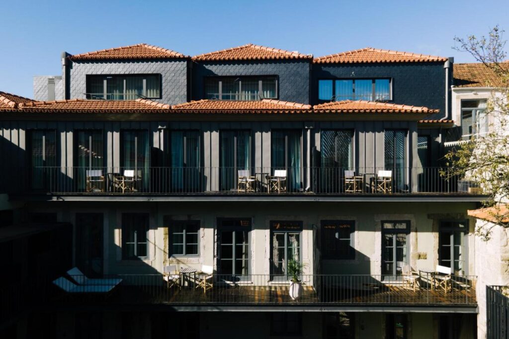 Top 12 Boutique Hotels in Porto, Portugal 26 A two-story building with dark roof tiles and exterior walls, featuring balconies on both floors with outdoor seating and tables—an inviting scene often found at boutique hotels in Porto under a clear blue sky.