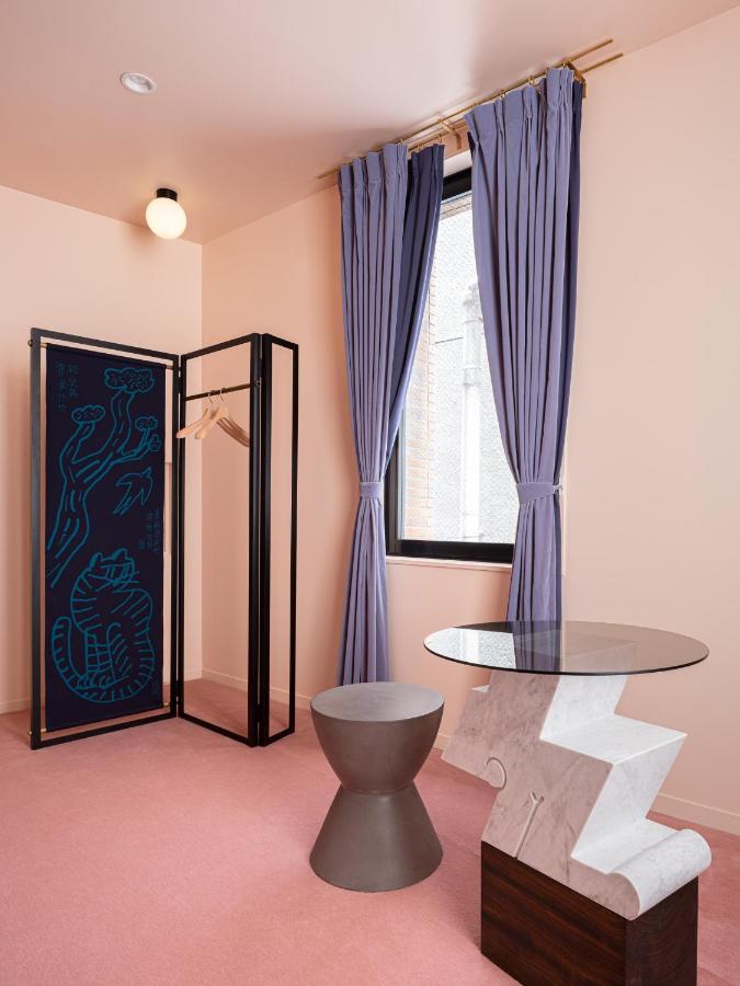 15 Unique Hotels in Tokyo: Coolest hotels 31 A stylish room in one of the unique hotels in Tokyo, featuring pink walls and floor, a window with lavender curtains, a black open wardrobe with a blue art panel, a gray stool, and a geometric marble and glass table.