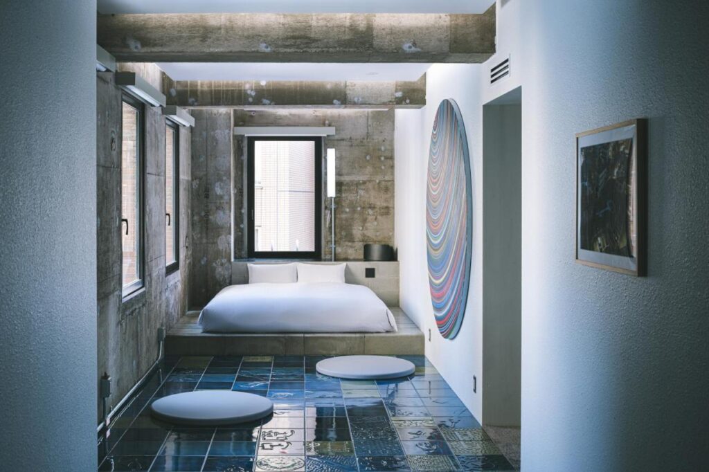 15 Unique Hotels in Tokyo: Coolest hotels 30 A modern, minimalist bedroom inspired by unique hotels in Tokyo, featuring a low platform bed, white bedding, tiled floors, large windows, exposed concrete walls, and colorful circular wall art beside the bed.