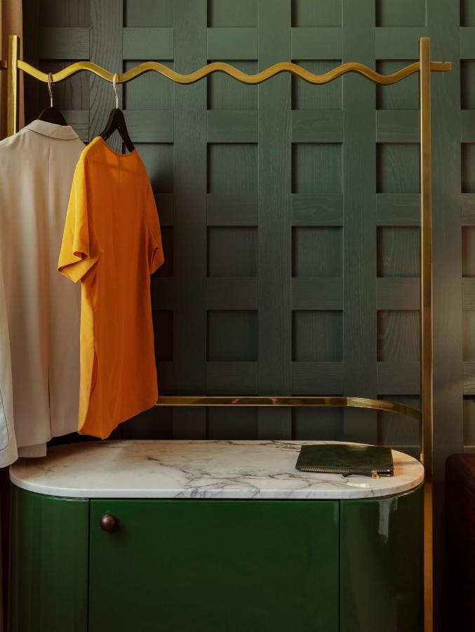 13 UNIQUE Boutique Hotels in Rome 26 A yellow T-shirt and a white shirt hang on black hangers from a gold clothing rack, evoking the chic style of boutique hotels in Rome, Italy. Below, a green and white marble-topped cabinet rests against a dark green grid-patterned wall.