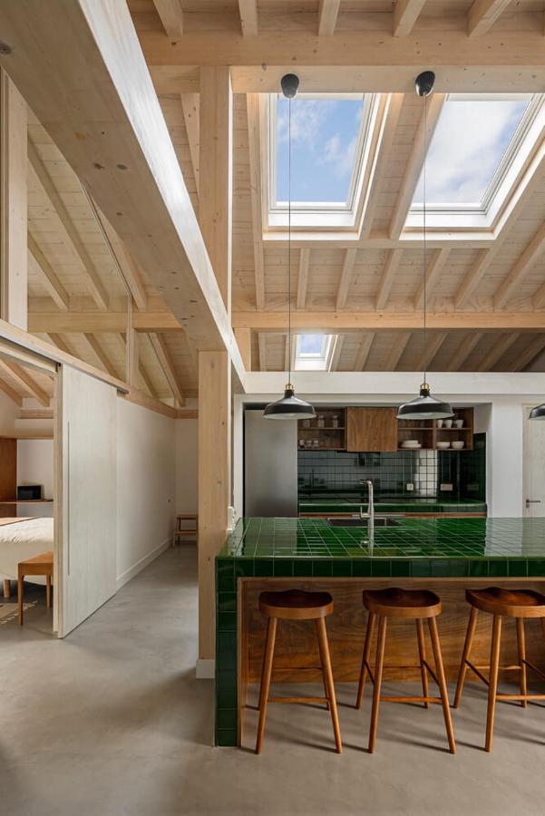 A modern kitchen with a green-tiled island, three wooden bar stools, pendant lights, wood ceiling beams, and large skylights—reminiscent of boutique hotels in Cantabria. An adjacent bedroom is visible through an open doorway on the left.