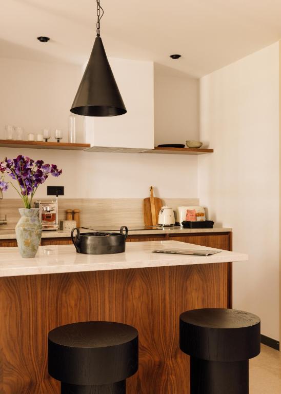 Top 12 Boutique Hotels in Porto, Portugal 34 Modern kitchen with a white countertop island, two black round stools, a black pendant light, wooden cabinets, open shelves with dishes, and a vase of purple flowers—reminiscent of the chic style found in boutique hotels in Porto.