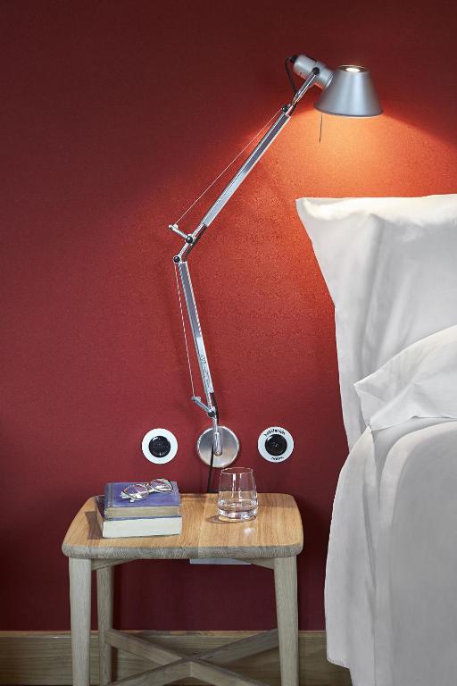 13 Unique Hotels in Barcelona, Spain 30 A wooden bedside table with books, a glass of water, and a modern adjustable desk lamp beside a bed with white bedding, set against a red wall—this cozy scene echoes the inviting style found in unique hotels in Barcelona.