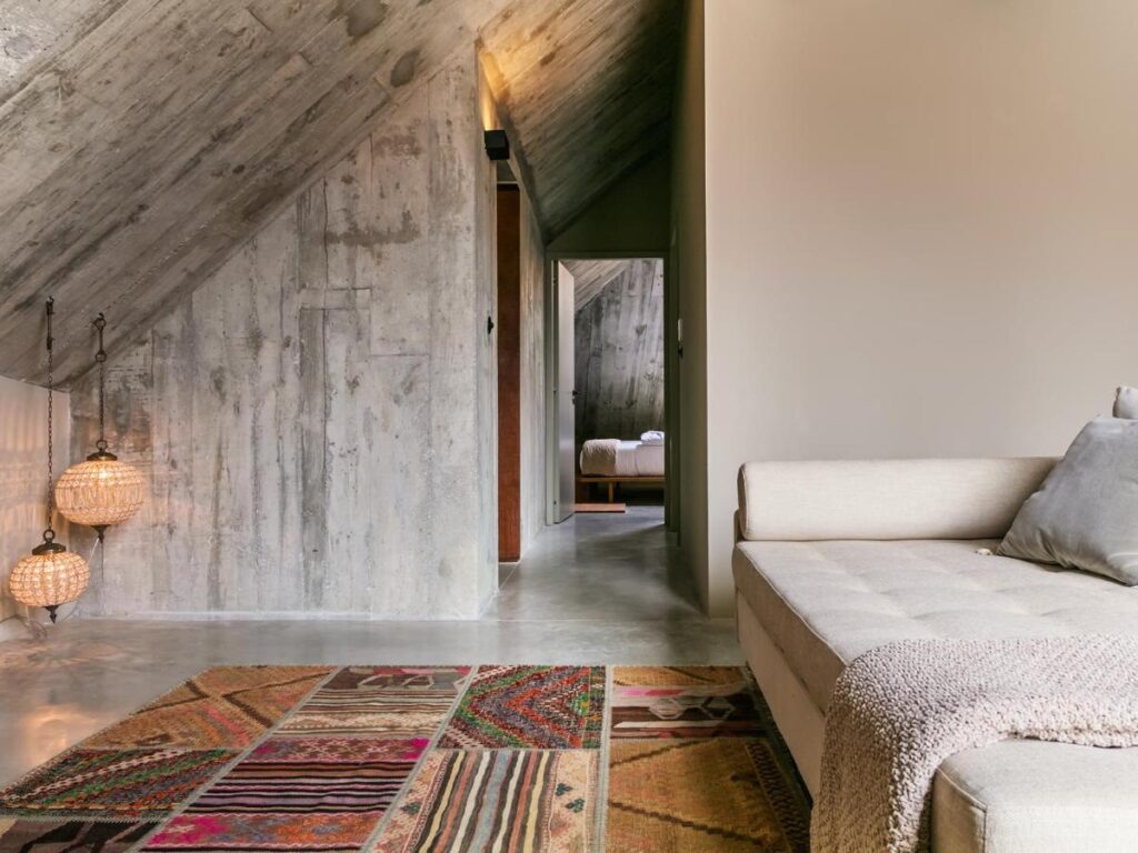 Top 12 Boutique Hotels in Porto, Portugal 23 A cozy attic room with concrete walls and ceiling, a beige sofa with cushions and a throw, colorful patchwork rug, hanging pendant lights—reminiscent of the charm found in boutique hotels in Porto—and a view into a bedroom with a neatly made bed.