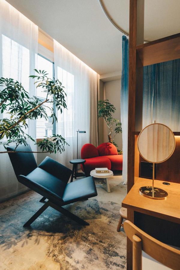15 Unique Hotels in Tokyo: Coolest hotels 13 A modern, cozy room in one of the unique hotels in Tokyo, featuring large windows with sheer white curtains, green potted plants, a black lounge chair, red loveseat, small round table with books, and a wooden vanity with a round mirror.